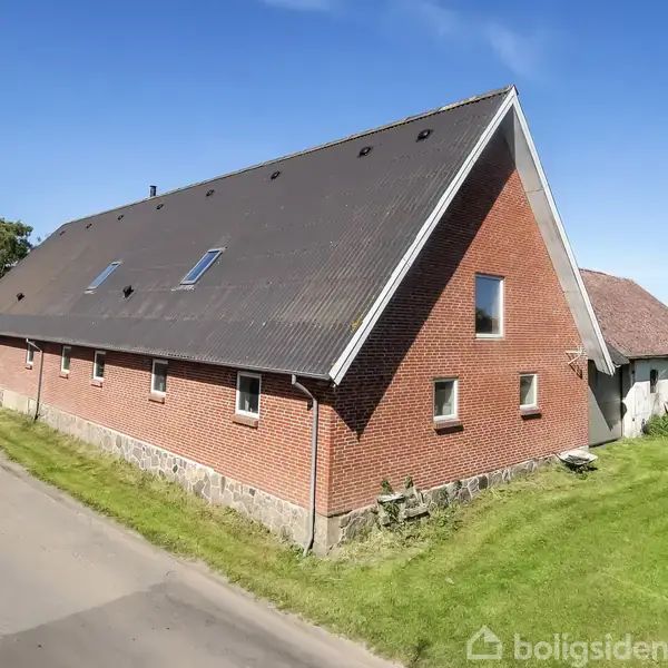 Stort rødt murstenshus med sort tag står ved vejen. Huset har flere vinduer og ligger på en græsplæne med træer og buske i nærheden under en klar blå himmel.