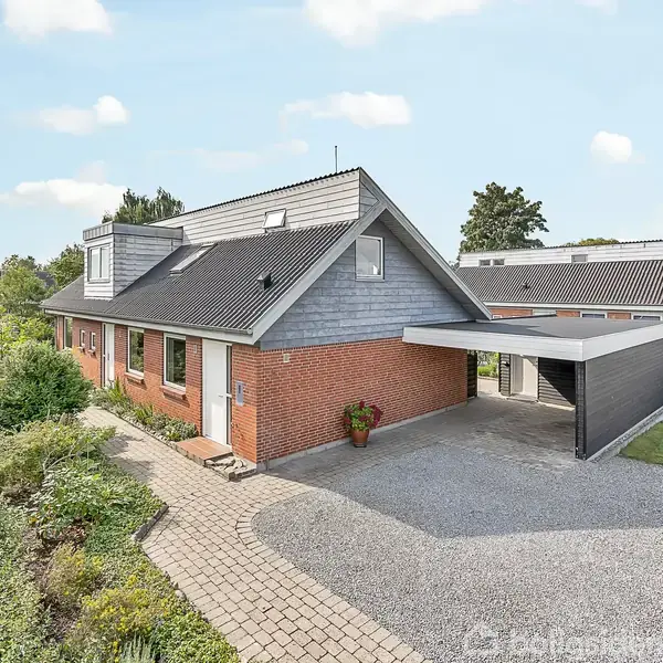 Et rødt murstenshus med gråt tag og carport står omgivet af en velplejet have og flisebelagt sti, under en klar blå himmel.