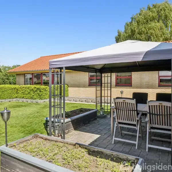 Pavillon med tag står over spisebord og stole på en terrasse i en have. Rød murstensbygning ses i baggrunden med blå himmel og grønne træer.