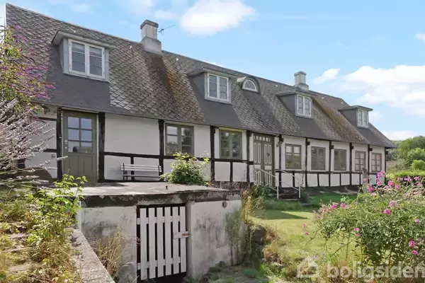 Et rækkehus med sort-hvide bindingsværk og flere vinduer ligger i en have med blomster og græs. Huset har en skrånende tagstruktur med flere kviste.