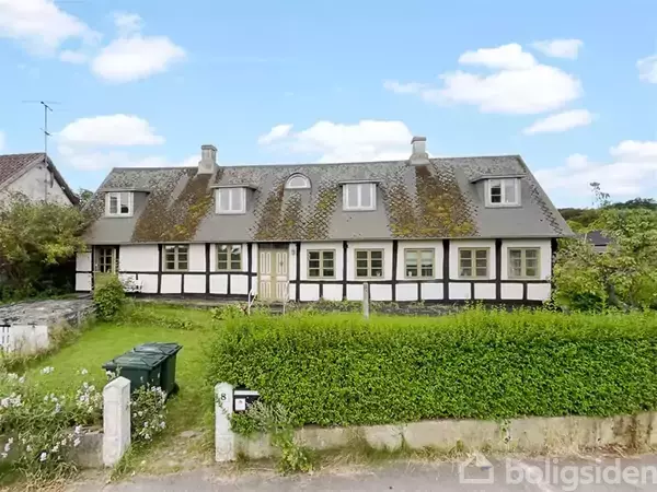 Et bindingsværkshus med mosbelagt tag står i en grøn have med hæk og en skraldespand ved fortovskanten. Klar blå himmel med spredte skyer i baggrunden.