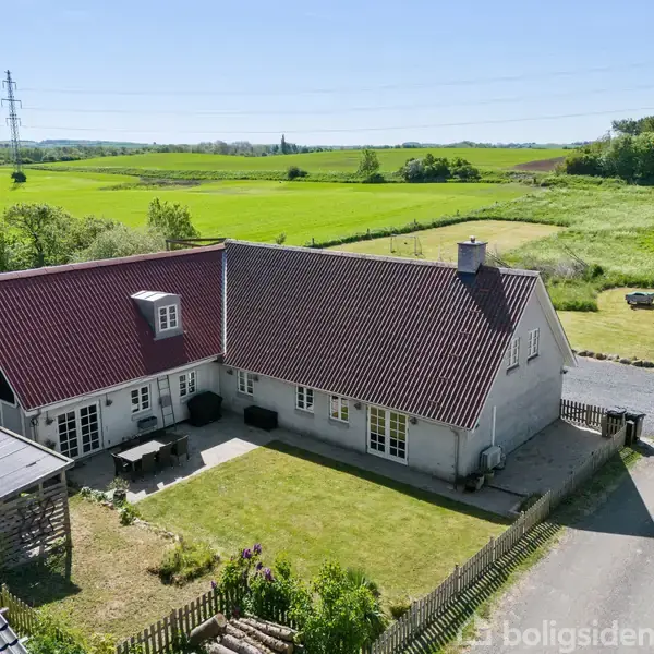 Et hvidt hus med rødt tag ligger i landlige omgivelser ved en vej. En lukket have med terrassemøbler ser ud til at byde på afslapning.