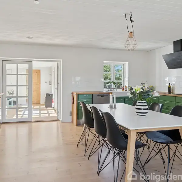 Et spisebord med sorte stole står i et rummeligt køkken med grønne skabe. En vase med blomster pryder bordet. Store vinduer og glasdøre giver rummet et lyst udtryk.