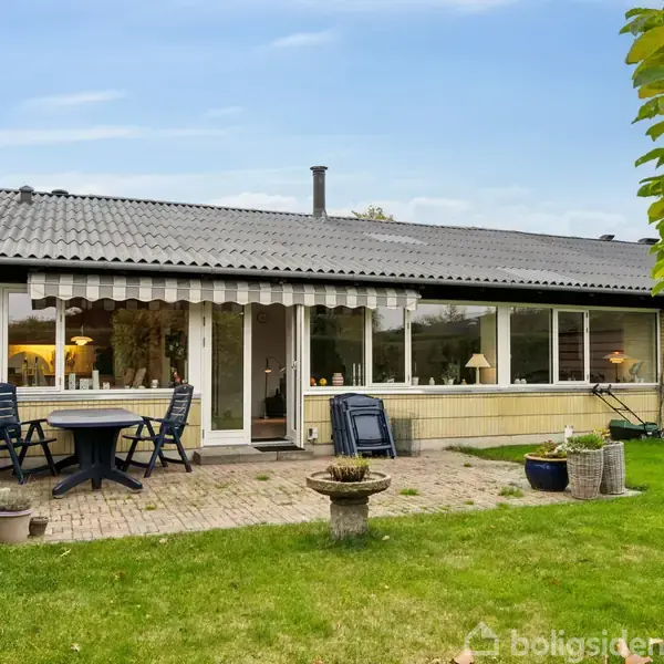 Et enfamiliehus med store vinduer står stille på en græsplæne, omgivet af grønne planter og en flisebelagt terrasse med havemøbler.