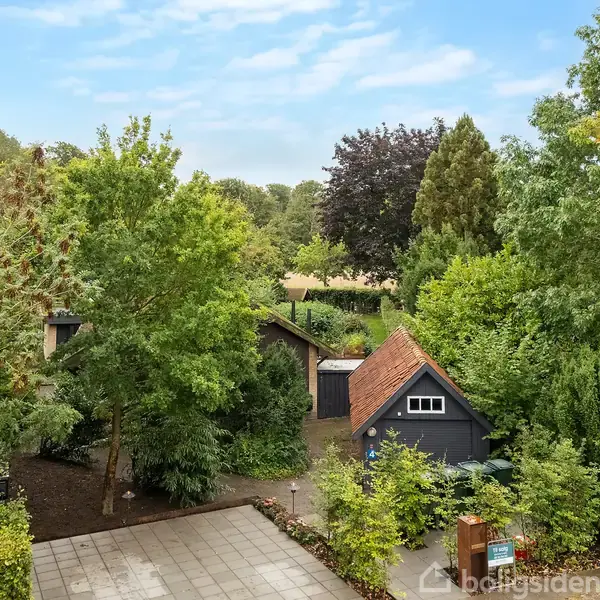 Hus med rødt tag ses blandt frodige træer og buske, beliggende i en grøn have. Omgivelserne er fyldt med tæt vegetation og stier, der skaber en naturrig atmosfære.