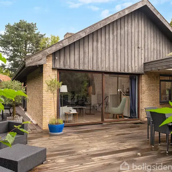 Et hus med træ- og murstensfacade har en stor terrasse med sort udendørsmøblering. Omkring terrassen er der grønne planter, træer samt blå himmel i baggrunden.