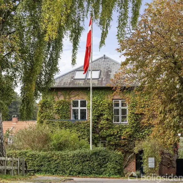 Hus dækket af klatrende planter, med en dansk flagstang foran. Omkranset af frodige træer, skaber en naturlig og rolig atmosfære omkring bygningen.