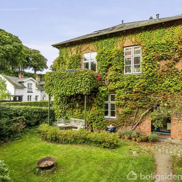 Murstenshus dækket af grønne planter, med en balkon med bænk. Beliggende i en frodig have med en græsplæne, buske og ildsted. Nabohuse og skov i baggrunden.