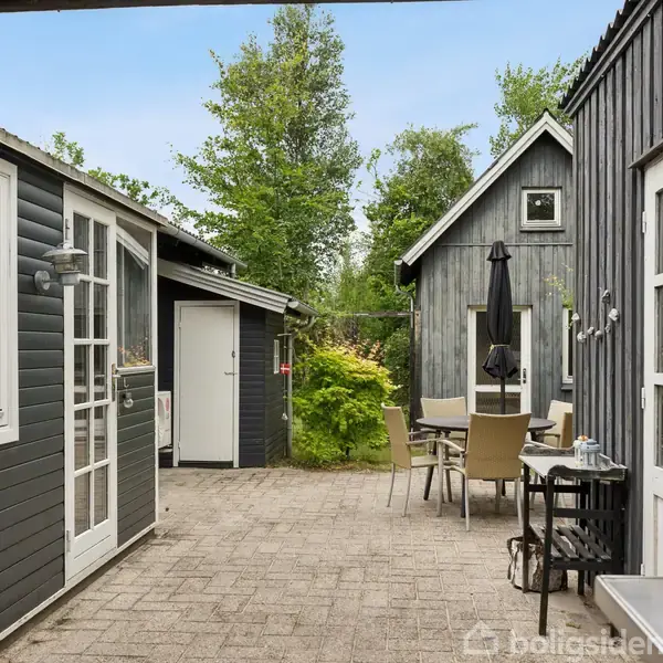 To sorte træbygninger omkranser en gårdhave med brostensbelægning. Fire stole og et bord med en parasol står i midten. Grønt løv omkranser området.