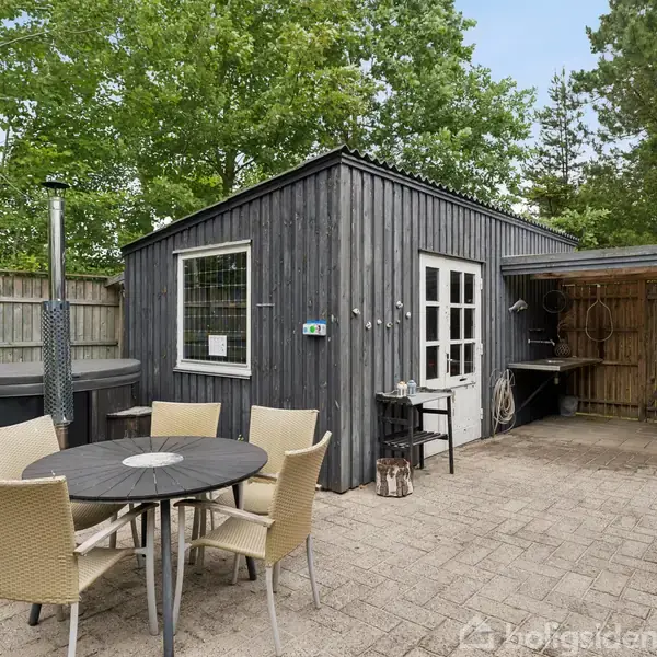 En rund bord med seks stole står på en terrasse foran et sort træskur med vinduer. Til venstre er et udendørs brændeovn. Omkring skuret er der et hegn og træer.