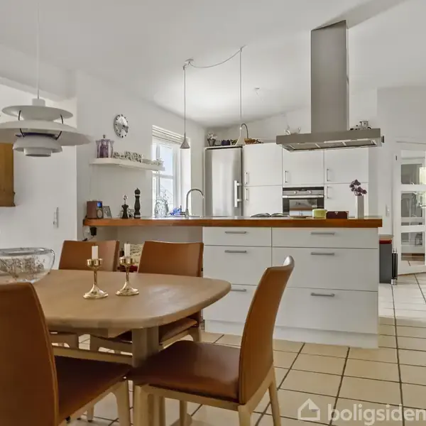Et moderne køkken med spisebord er omgivet af brune stole. Køkkenet har hvide skabe og en central kogeø med emhætte. Rummet er lyst med fliser på gulvet.