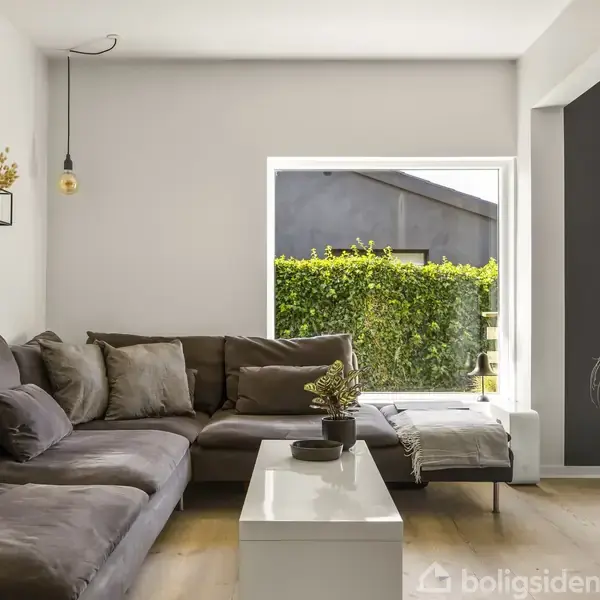 En grå hjørnesofa står i en stue ved siden af et stort vindue, som giver udsigt til en grøn hæk udenfor. En hvid reol står foran sofaen.