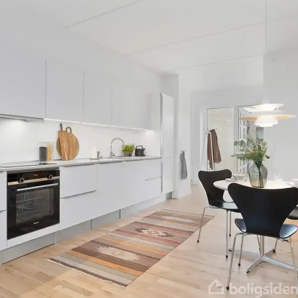 Et moderne køkken med hvide skabe og indbygget ovn. Bordet har sorte stole og en vase med blomster. Tæppe på trægulvet. Lysekrone ovenover, med lys indretning.