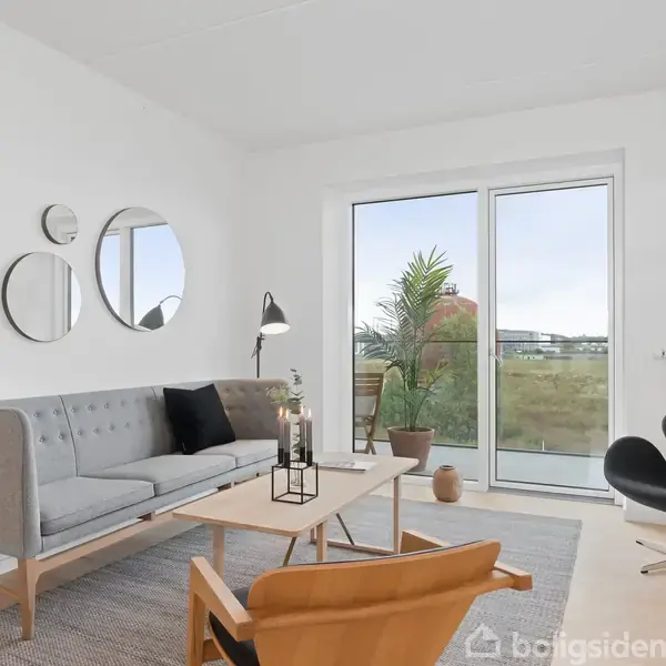 En grå sofa står i et lyst stuehjørne med to runde spejle på væggen. En lænestol og en rund stol omkranser et sofabord. Store vinduer fører ud til en altan.