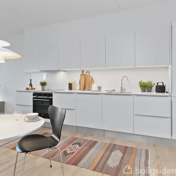 Et moderne køkken med hvide skabe langs en væg; planter, skærebrætter og en kedel står på bordpladen. Et rundt spisebord med sort stol i forgrunden, belysning ovenover.
