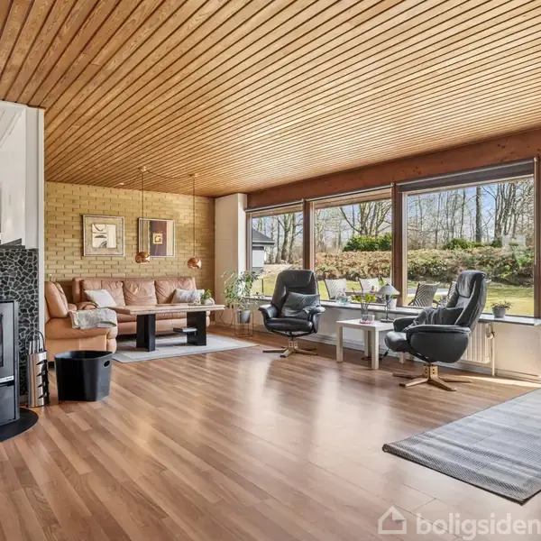 Brændeovn opvarmer stue med træloft og gulv. Store vinduer giver udsigt til en have. Stueindretningen inkluderer en sofagruppe og skrivebord med kontorstole.