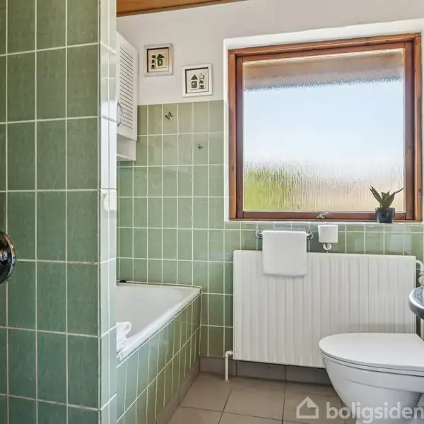 Badeværelse med grønne fliser omkring badekar og bruser. Et vindue over radiator med plante ved siden af. Toilet, spejl og håndvask på højre side.