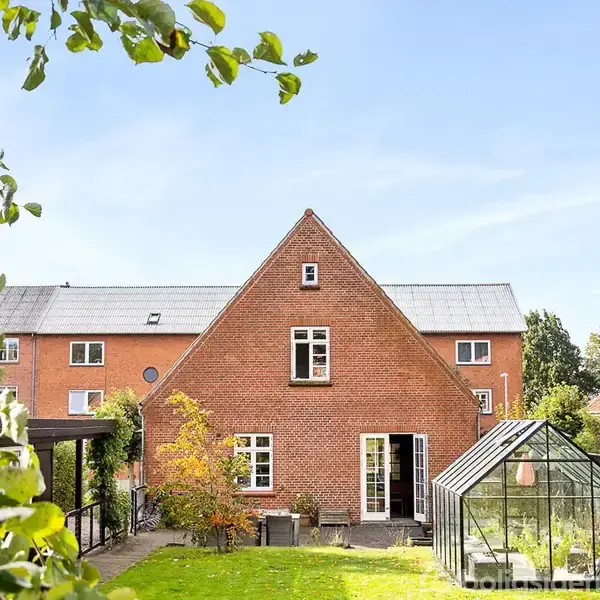 Rødt murstenshus står i solen med en lille have. Et drivhus står til højre, træer med æbler omkranser stedet, og himlen er klar blå.