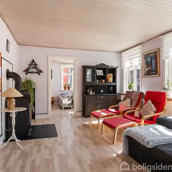 Stue med to røde stole og sort sofa. Et lille bord med lampe ved siden af en sort brændeovn. Sollys strømmer ind fra vinduerne på højre side.