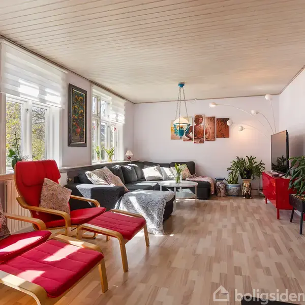 Stue fyldt med møbler; to røde lænestole og en stor grå sofa set mod lyse trævægge og -gulve, dekoreret med malerier, planter og lamper, i dagslys.