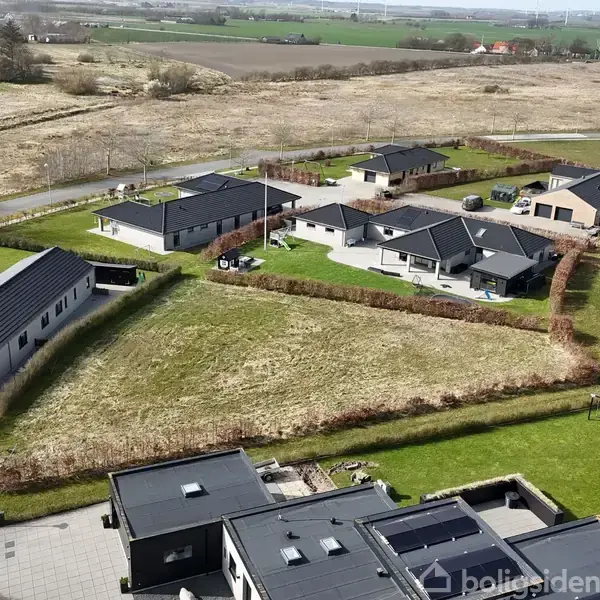 Luftfoto viser et moderne boligområde. Huse med sorte tage omgivet af velplejede græsplæner ligger ved siden af hinanden. I baggrunden ses åbne marker under en klar himmel.