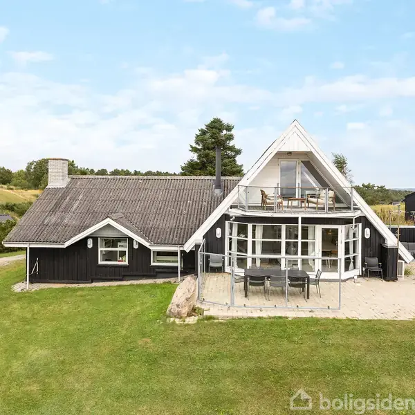 Sort sommerhus med store vinduespartier og terrasse, står på en stor græsplæne. Huset har en markant A-formet tagetage. Omgivet af natur og andre huse i baggrunden.