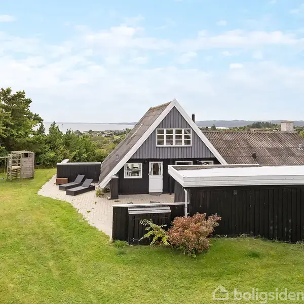 Et sort træhus står på en græsplæne med træer i baggrunden. Huset har et spidst tag og en terrasse med havemøbler. Udsigt til skov og hav i horisonten.