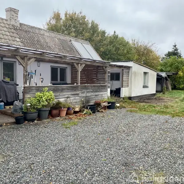 Et hus med veranda er omgivet af grusbelagt gårdsplads. Krukker med planter står langs væggen. Bag huset er der træer og grønne områder, som skaber en landlig atmosfære.