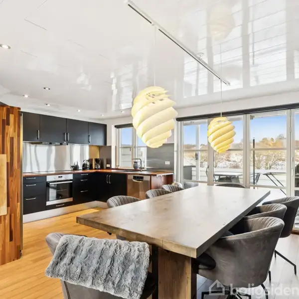 Et moderne køkken-alrum med træbord, omgivet af polstrede stole med gråt overtæk. Store vinduer giver lysindfald fra terrassen. To spiralformede lamper hænger over bordet.