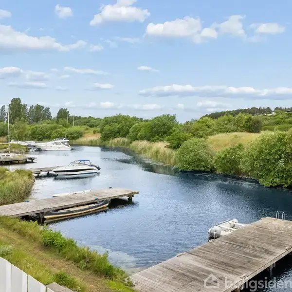 En båd ligger fortøjet ved en smal, rolig kanal omgivet af grønne buske og græs. Flere både er til stede, og moderne huse kan ses i baggrunden under en skyet himmel.