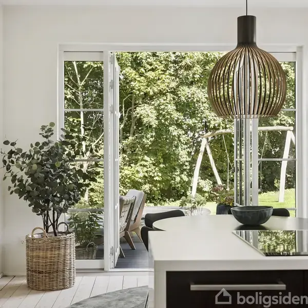 Lampen hænger over et køkkenbord, mens terrassedøre står åbne mod en have med træer og en gynge. En plante i en kurv står ved siden af dørene.