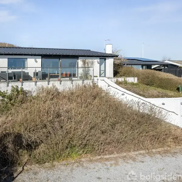 Et moderne hus med store vinduer står på en skråning omgivet af buske. En garage ligger til højre under en hvid trappe, der fører op til indgangen.