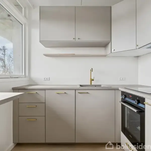 Køkken med grå skabe og skuffer, integreret sort ovn og moderne vandhane. Store vinduer til venstre giver naturligt lys. Rent og minimalistisk design med lys trægulv.
