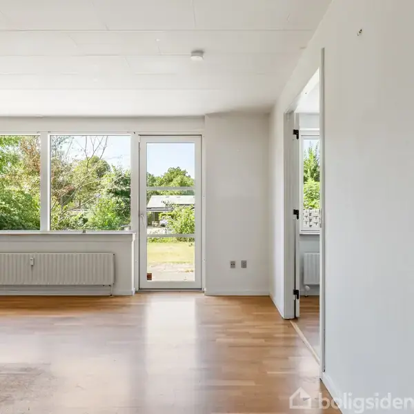 Et lyst rum med trægulv og store vinduer ud mod en have. En åben dør fører til en udendørs terrasse. Radiator under vinduet.