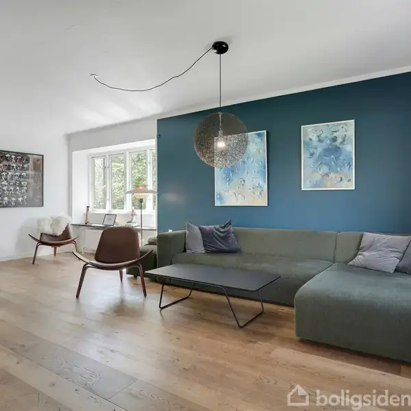 En stue med en stor hjørnesofa placeret ved en blå væg, udsmykket med to abstrakte malerier. Rummet har lyse trægulve, moderne stole, og et rundt loftslys.