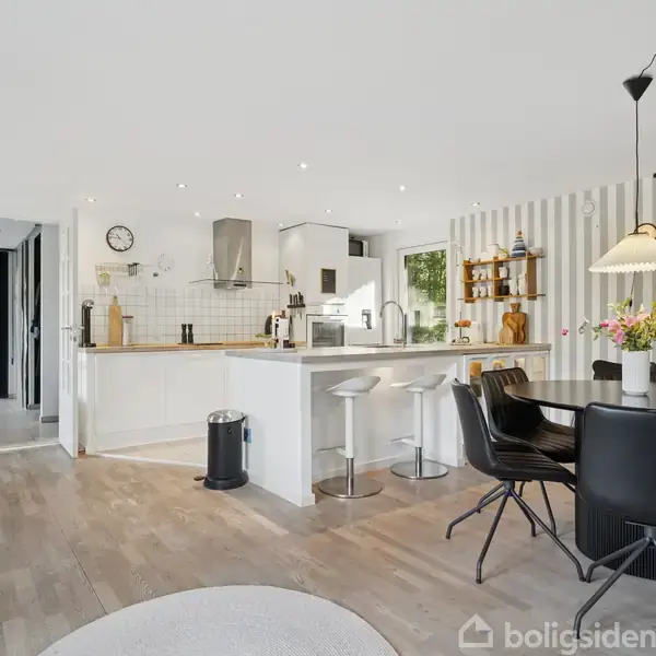 Køkkenet har en hvid køkkenø med to barstole. Ved siden af er der spisebord med sorte stole og blomster i en vase. Rummet er lyst og moderne indrettet.