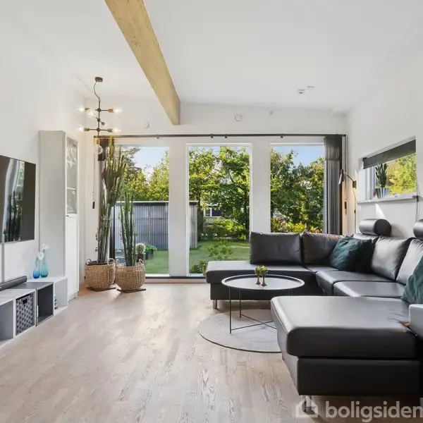 Stor sort lædersofa står foran et stort vindue, der viser en grøn have. Et moderne TV hænger på væggen, og rummet er lyst med dekorativt interiør og træelementer.