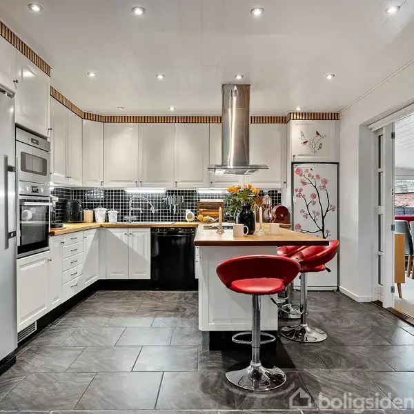 Et moderne køkken med hvidt inventar og sort flisegulv. To røde barstole står ved en køkkenø. Blomster og kunst dekorerer rummet. Døre fører til andre rum.