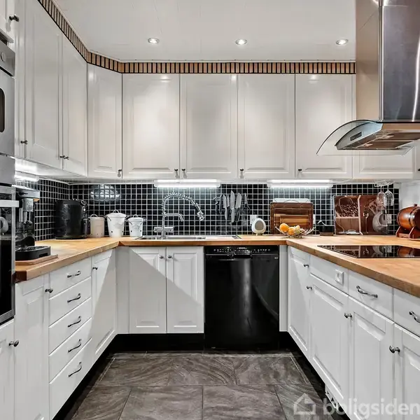 Et moderne køkken med hvide skabe, en indbygget ovn og sort flisebagplade. En vase med orange blomster står på bordpladen. Området er veloplyst med indbyggede loftlamper.