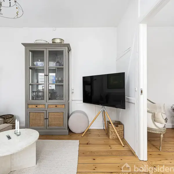 Et moderne stueinteriør med en grå vitrineskab står mod væggen. Et tv på trefod står til højre. En beige sofa og et rundt sofabord er placeret til venstre.