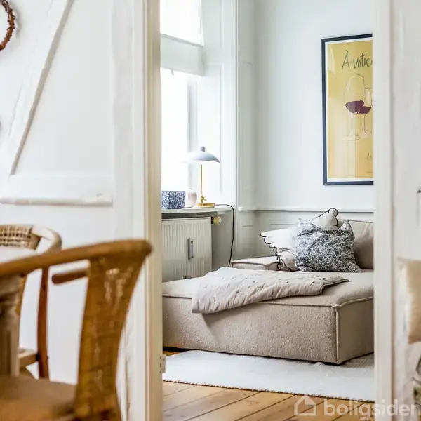 En lille, hyggelig stue med en beige sofa pyntet med puder og tæppe. Ved siden af er et bord med en lampe. Stuen er dekoreret med vægkunst og kurvemøbler.