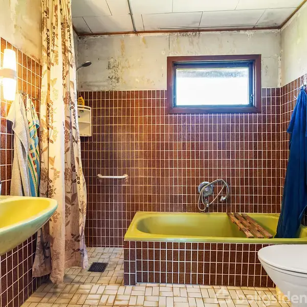 Et badeværelse med brune fliser. En gul vask og badekar. Vinduet er placeret højt, gardin hænger ved badet. Toilettet står tæt på badekarret. Lyft i siden.