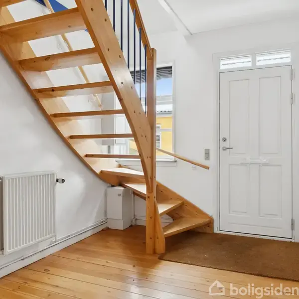 Trætrappe fører opad i en lys gang med trægulv. Til højre ses en hvid indgangsdør og til venstre et radiator under et vindue.