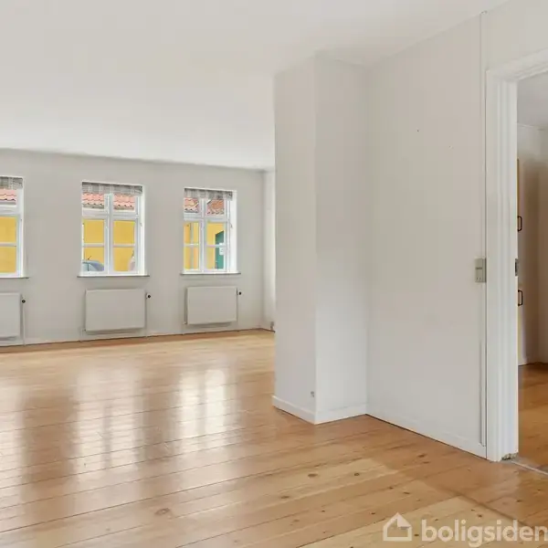 Et lyst rum med trægulv, en åben dør til venstre viser en garderobe, mens et vindue til højre slipper lys ind. Hvide vægge skaber en ren atmosfære.