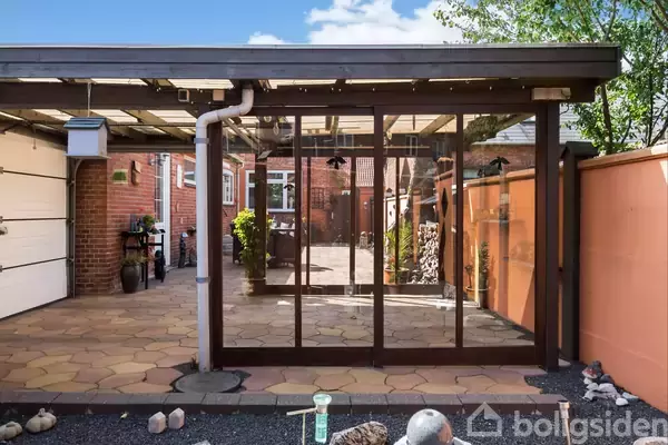 Pergola med glasvægge står i en indhegnet gårdhave. Murstensbygninger danner baggrunden, og gulvbelægningen er af betonsten. Dekorative sten ligger langs kanten.