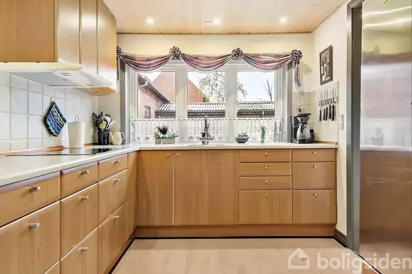 Køkken med lyse træskabe og skuffer langs væggene. Vindue i enden med lilla gardiner, kaffemaskine og køkkenredskaber placeret på bordet. Flisebagvæg og lyst træloft.