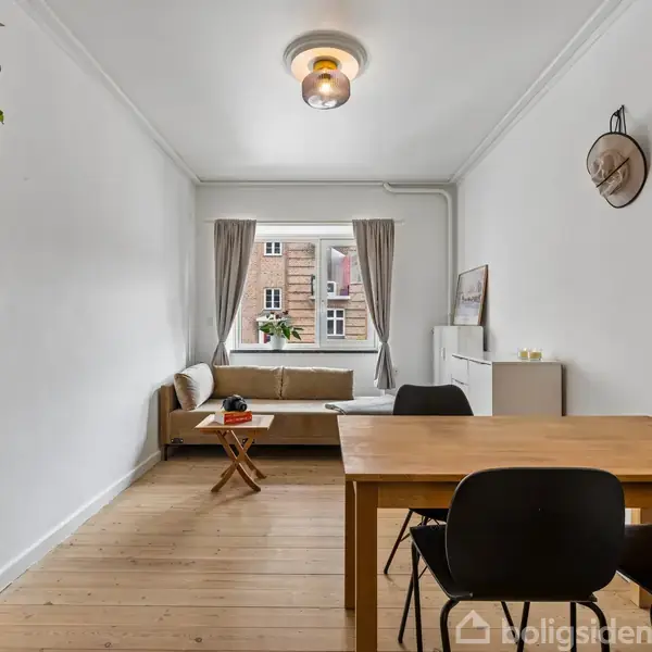 Stue med spisebord og to stole, sofa ved vindue, hylde med bøger til venstre. Omgivet af lyse vægge, trægulv og en loftlampe.