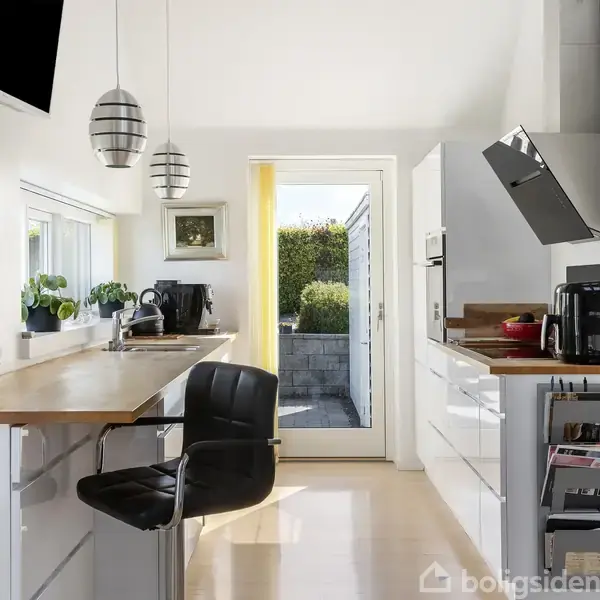 Et moderne køkken med træbordplade og sort læderstol. Køkkenet har hvide skabe og stålapparater under pendellamper. En glasdør leder til en udendørs gårdhave omkranset af hæk.