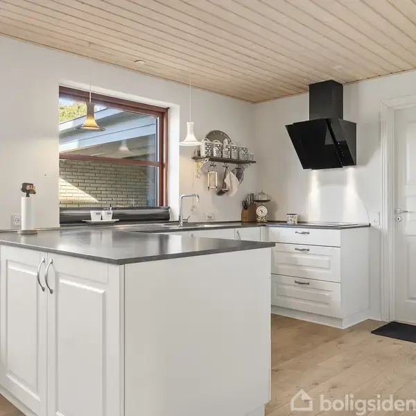 Et moderne køkken med hvidt køkkenbord står centralt. Skinnende grå bordplade pryder overfladen. Baggrunden viser lyse vægge, store vinduer og en træbeklædt loft med en emhætte.