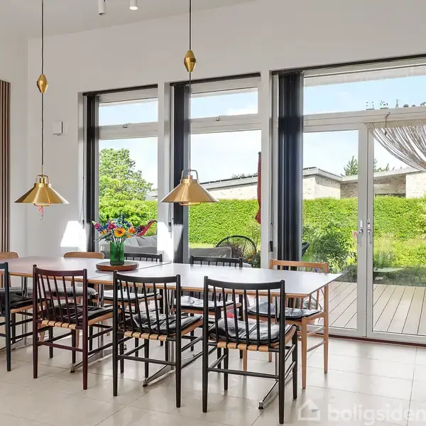 Et spisebord med otte stole står i et lyst rum. Tre hængelamper hænger over bordet. Store vinduer og en glasdør fører ud til en terrasse med grøn udsigt.